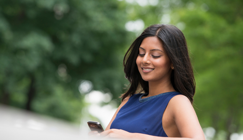 A woman with glowing skin sits outside and smiles as looks at her phone